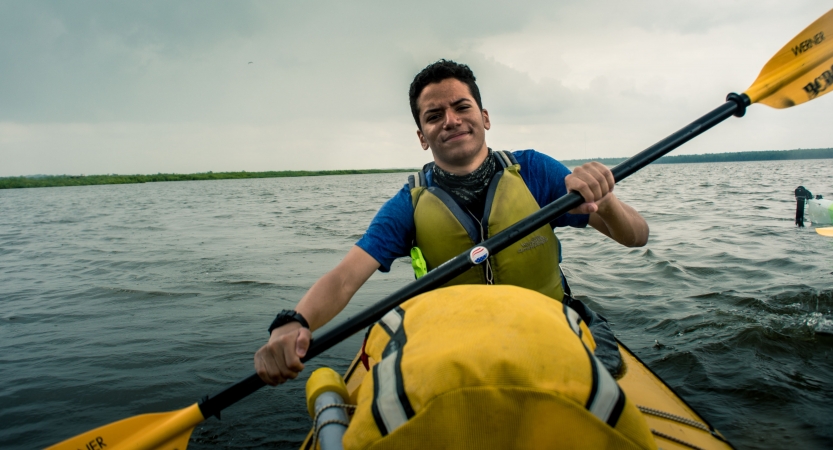 a young person paddling a yellow kayak smiles at the camera. There is a vast body of water behind them. 